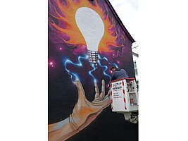 Eine Hauswand mit einem grossflächigen Graffiti. Der Künstler steht auf einem Kran um dieses zu bearbeiten.