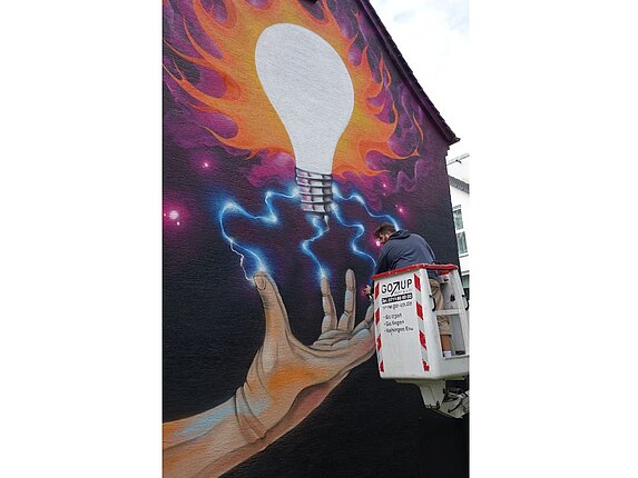 Eine Hauswand mit einem grossflächigen Graffiti. Der Künstler steht auf einem Kran um dieses zu bearbeiten.