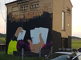 Das technische Gebäude in Darmsheim bekommt ein eigenes großflächiges Graffiti. Die ersten bunten Flächen und Motive sind zu erkennen - Hände, weisse Spielkarten, schwarzer Hintergrund.