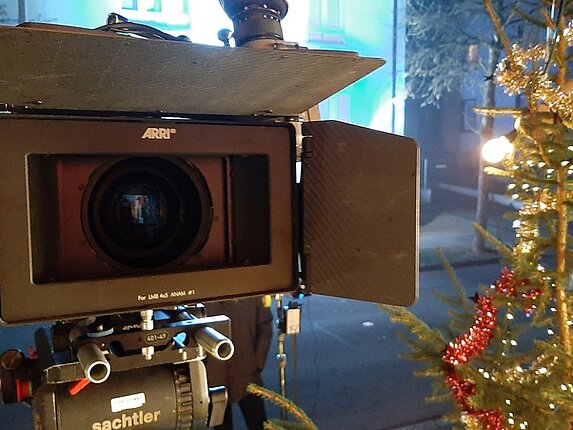 Nahaufnahme einer Filmkamera von vorn (Linse). Am Rechten Rand ist ragt ein geschmückter Weihnachtsbaum ins Bild.