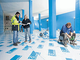 Bauarbeiter und Projektleiter stehen und arbeiten in einer mit Säulen versehenen Halle. Boden und Wände sind mit Folien geschützt.