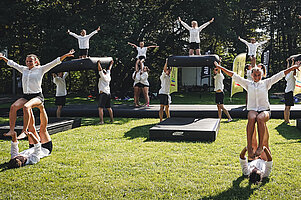 Tanzperformance vieler Cheerleader in weiß-schwarzer Kleidung auf einer Wiese. Einige von ihnen performen eine Hebefigur, zwischen Ihnen liegen schwarze Gymnastikmatten.