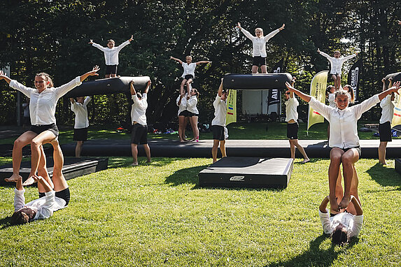 Tanzperformance vieler Cheerleader in weiß-schwarzer Kleidung auf einer Wiese. Einige von ihnen performen eine Hebefigur, zwischen Ihnen liegen schwarze Gymnastikmatten.