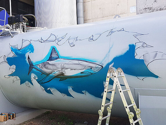 Erste Skizzen und Umrisse für das großflächige Graffiti sind auf einem Öltank gezeichnet.