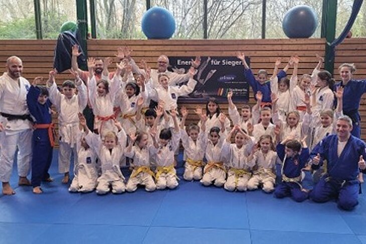Gruppenbild einiger Kinder und Jugendlicher mit Judo-Trainingskleidung in der Turnhalle. Alle freuen sich und strecken die Hände in die Luft.