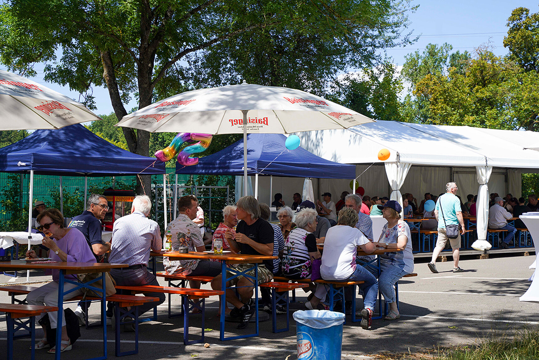 Einige Menschen sitzen an Biertischgarnituren unter Sonnenschirmen und essen einen Snack.
