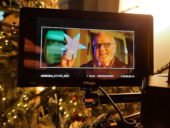 Nahaufnahme des Displays einer Filmkamera. Dort zu sehen ist Herr Hoffmann, lächelnd mit einem Weihnachtsstern in der Hand. Im Hintergrund des eigentlichen Bildes steht ein geschmückter Weihnachtsbaum.