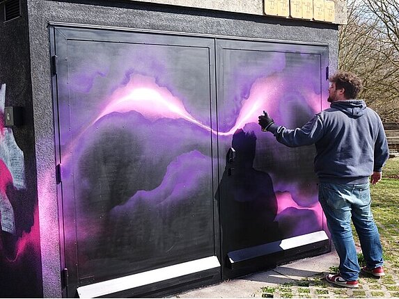 Ein technisches Gebäudes wird mit einem Graffiti verschönert. Der Künstler sprüht rosafarbene Blitze auf schwarzen Grund an die Hauswand.