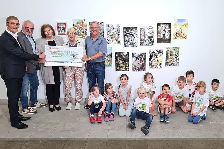 Herr Hoffmann übergibt den Spendenscheck an den Verein kids@kita. Die Gruppe Erwachsener steht vor einer mit vielen Zeichnungen behangenen Wand. Einige Kinder sitzen vor der Wand auf dem Boden.