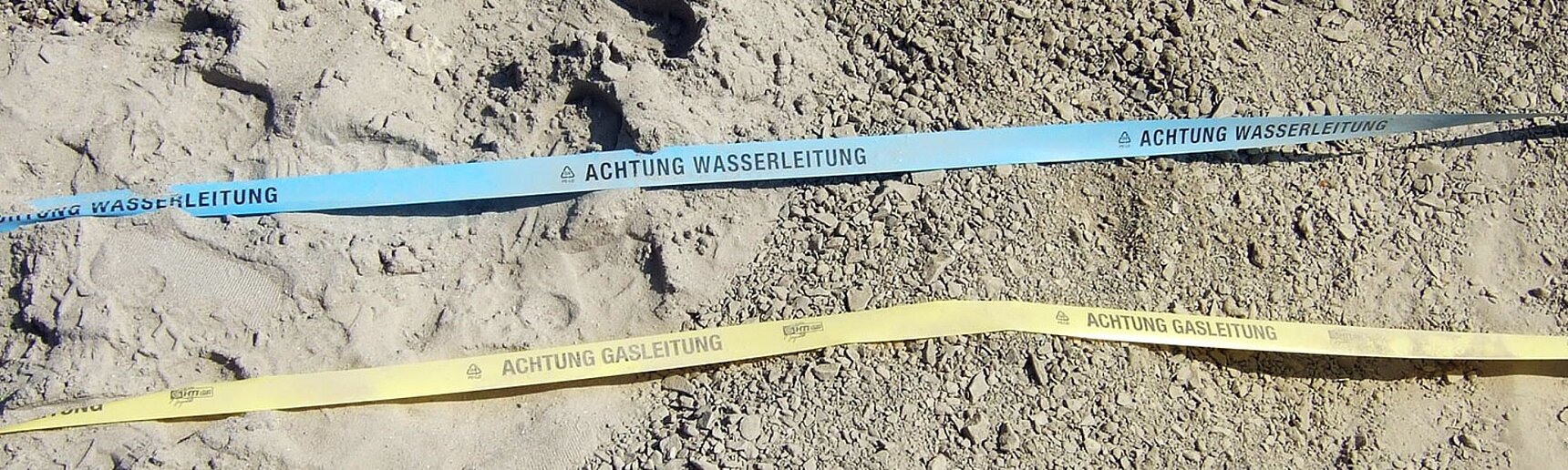 Ein blaues Band mit der Aufschrift "Achtung Wasserleitung" und ein gelbes Band mit der Aufschrift "Achtung Gasleitung" liegen auf einem schotterigen Boden.