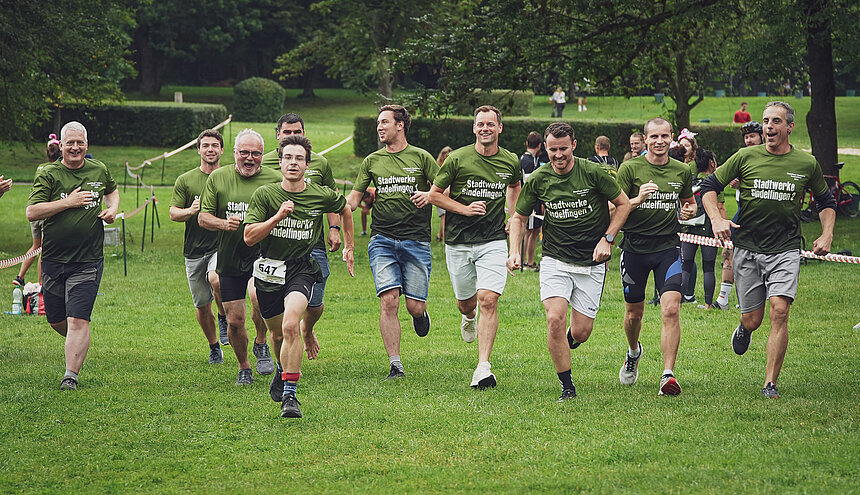 Mehrere Personen in Teamkleidung rennen freudig auf einer Wiese dem Betrachter entgegen.