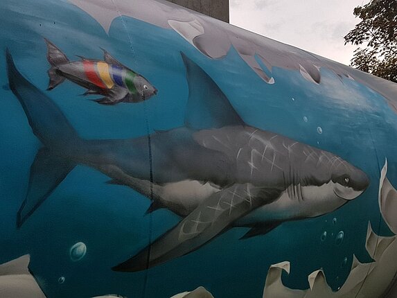 Ein Graffiti auf einem Öltank. Das Motiv: Es scheint als wäre da ein riesiger Riss im Tank, die Ränder sind ausgefranst und mann kann ins Ineere schauen. Dort schwimmt ein grinsender Hai und ein kleiner Fisch in den Farben der Stadtwerke Sindelfingen in hellblauem Wasser.