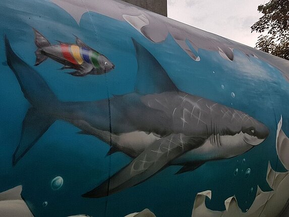 Ein Graffiti auf einem Öltank. Das Motiv: Es scheint als wäre da ein riesiger Riss im Tank, die Ränder sind ausgefranst und mann kann ins Ineere schauen. Dort schwimmt ein grinsender Hai und ein kleiner Fisch in den Farben der Stadtwerke Sindelfingen in hellblauem Wasser. 