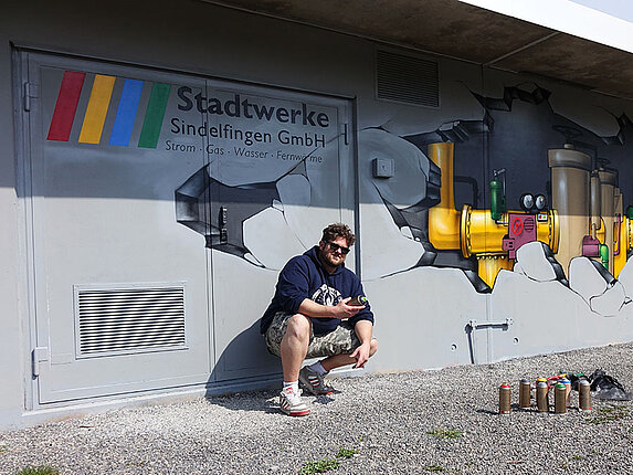 Gasdruckregelstation in der Rudolf-Harbig-Straße Der Graffiti-Künstler hockt vor seinem Werk. Er hat eine Spraydose in der hand, weitere Dosen stehen vor ihm auf dem Boden. An der Hauswand erkennt man das Logo der Stadtwerke Sindelfingen, sowie das Motiv der aufgebrochenen Hauswand und der darin befindlichen Bauarbeitern.