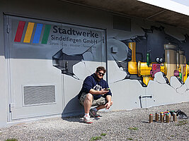 Der Graffiti-Künstler hockt vor seinem Werk. Er hat eine Spraydose in der hand, weitere Dosen stehen vor ihm auf dem Boden. An der Hauswand erkennt man das Logo der Stadtwerke Sindelfingen, sowie das Motiv der aufgebrochenen Hauswand und der darin befindlichen Bauarbeitern.