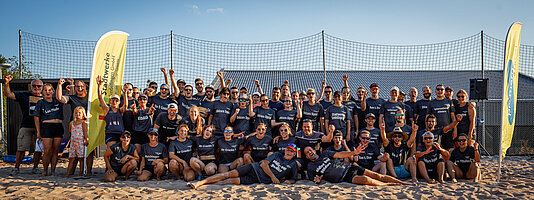 Gruppenbild vieler freudiger Teilnehmer auf einem sandigen Volleyballfeld. Viele sind barfuß und haben eine Sonnenbrille auf.