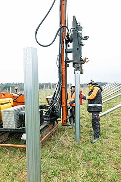 Zwei Bauarbeiter bringen mit einem schweren Baufahrzeug große Metallstangen in den Boden.