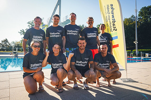 Teamfoto einiger Personen, barfuß oder in Badelatschen, am Rand eines Schwimmbeckens. Strahlend blauer Himmel, Sonnenschein und am rechten Rand steht ein Banner der Stadtwerke Sindelfingen.