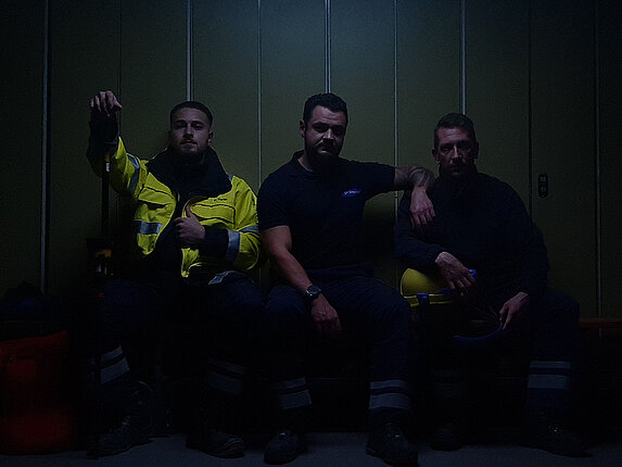 Sehr dunkle Szenerie in einer Umkleide. Drei Mitarbeiter in Montur sitzen auf einer Bank und schauen direkt zum Betrachter.