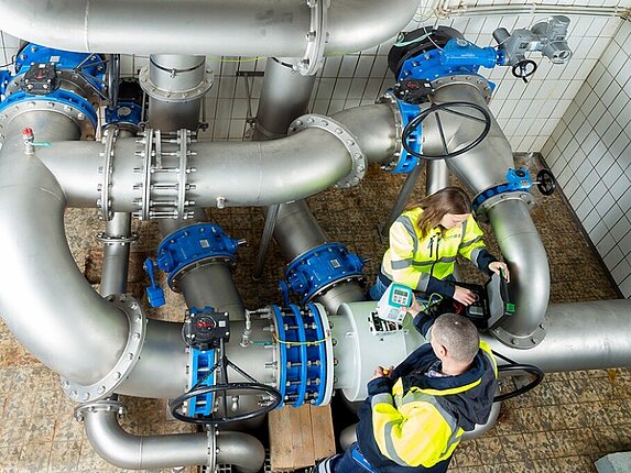 Kontrollen im Hochbehälter Buchen Zwei Mitarbeiter stehen inmitten von dicken Wasserrohren und untersuchen diese.