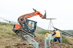 Ein Baggerfahrer und zwei Bauarbeiter setzen eine Betonplatte zwischen Metallpfosten. Im Hintergrund steht die bisher entstandene Fotovoltaikanlage.