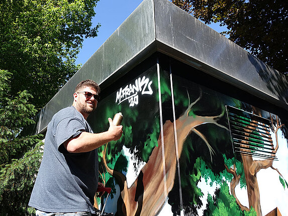 Der Graffiti-Künstler taggt sein Kunstwerk. Er schaut direkt in die Kamera und hebt den Daumen.
