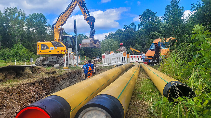 Mehrere Bauarbeiter, Bagger und Baufahrzeuge an einer Baustelle. zwei Arbeiter stehen in einem graben, daneben liegen dicke Rohre auf der Wiese.