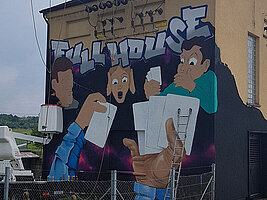 Das technische Gebäude in Darmsheim bekommt ein eigenes großflächiges Graffiti. Mehrere Personen, Hände, weisse Spielkarten und die Worte "Full House" sind zu erkennen.