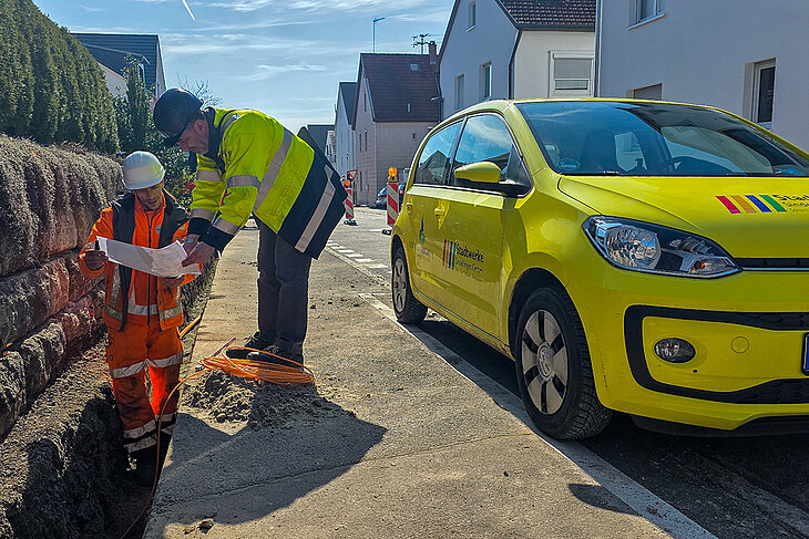 Zwei Mitarbeiter der Stadtwerke arbeiten an der Verlegung von Glasfaserkabeln in einem Graben. Daneben steht ein Auto der Stadwerke