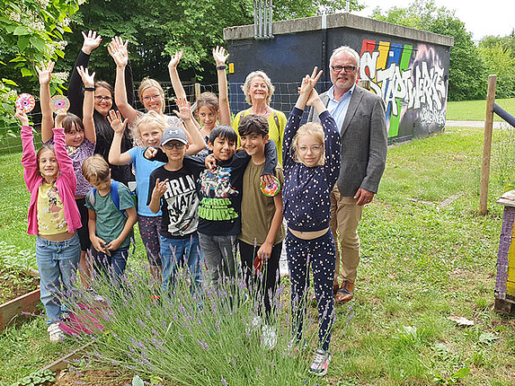 Eine Gruppe Kinder und Herr Hoffmann sind im Schulgarten "Gartenprojekt". Die Kinder heben die Hände in die Luft und freuen sich.