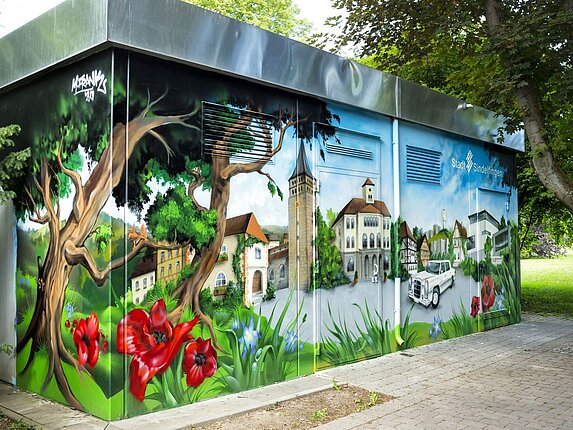 Ein technisches Gebäude mit einem grossflächigen Graffiti. Das Motiv: Der Marktplatz von Sindelfingen, umrahmt von grossen Bäumen und Blumen.
