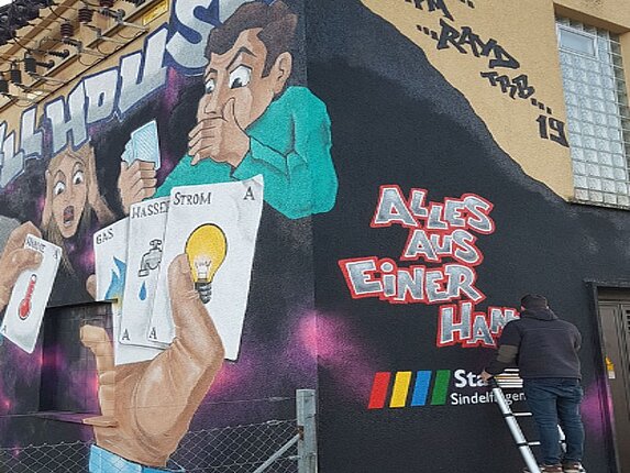 Schaltanlage zwischen Darmsheim und Ehningen Seitenansicht des technischen Gebäudes in Darmsheim mit einem großflächigen Graffiti einer Pokerrunde. Dort stehen die Worte "Alles aus einer Hand" und das Logo der Stadtwerke Sindelfingen. Der Künstler steht auf einer Leiter, die an der Hauswand lehnt.
