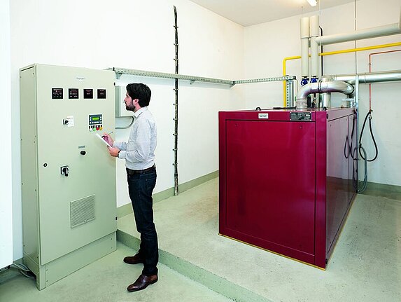 Ein heller Technikraum mit einem rotem Blockheizkraftwerk für die Strom- und Wärmeerzeugung. Ein Mitarbeiter steht an einem Schlatschrank und notiert sich einige Messwerte, die auf der Anzege angezeigt werden. Es verlaufen nur wenige Leitungen oder Rohre an den Wänden oder der Decke.