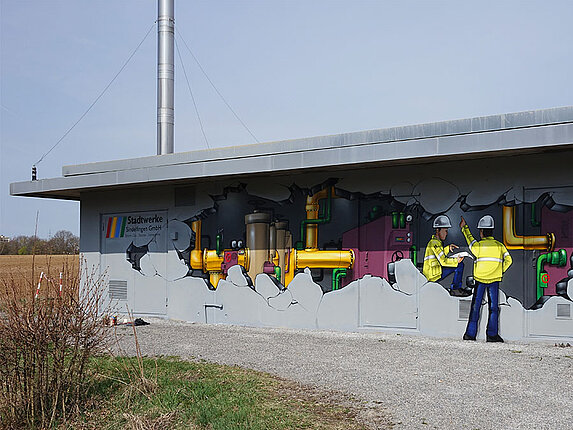 Gasdruckregelstation in der Rudolf-Harbig-Straße Motiv des Graffitis: Es scheint als wäre die Hauswand aufgebrochen und man können in das Innere des Gebäudes schauen. Zwei Bauarbeiter in gelber Sicherheitsjacke und mit Bauhelm befinden sich zwischen vielen Rohren und technischen Anlagen.