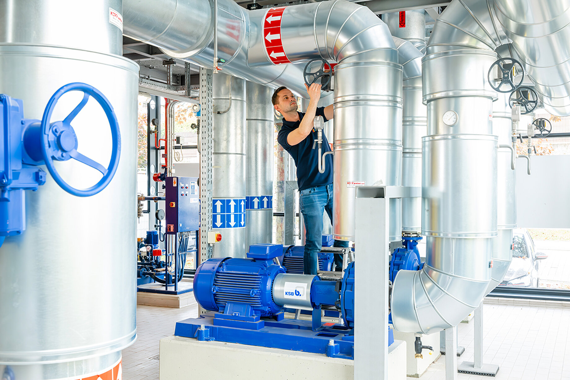 Ein heller Raum voller silberner Röhren und einige kleinere Turbinen. Ein Mitarbeiter kontrolliert dort die Fernwärmeversorgung. 
