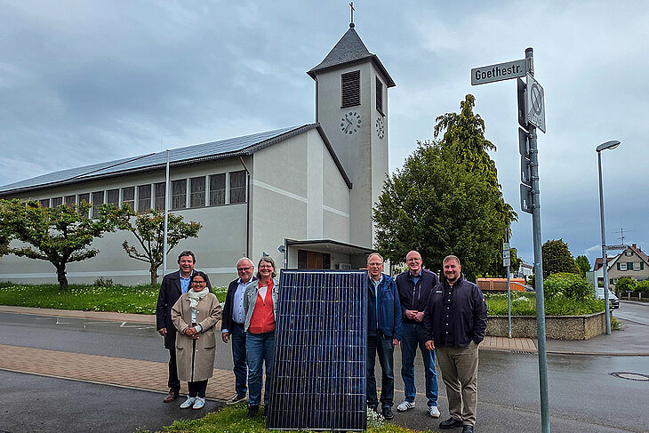 Vertreter/-innen der Kirche, der Stadt Gärtringen und der Stadtwerke Sindelfingen stehen neben einem PV-Modul. Im Hintergrund zu sehen ist die katholische Kirche Gärtringen mit der PV-Anlage auf dem Dach