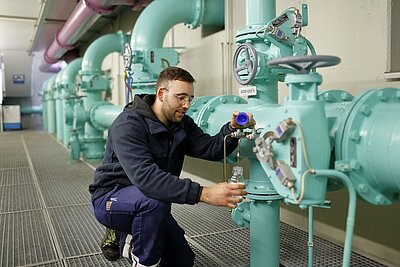 Ein Mitarbeiter hockt in einem Gang neben einem großen Wasserrohr und entnimmt eine Wasserprobe.