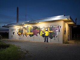 Nahaufnahme eines Graffitis bei Nacht: Es scheint als wäre die Hauswand aufgebrochen und man können in das Innere des Gebäudes schauen.Zwei Baurbeiter in gelber Sicherheitsjacke und mit Bauhelm befinden sich zwischen vielen Rohren und technischen Anlagen.