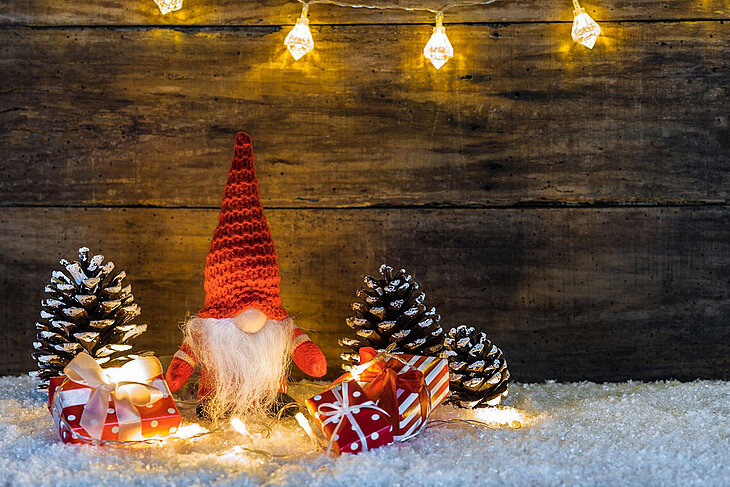 Eine kleine Wichtelfigur steht zwischen Tannenzapfen und einer Lichterkette auf einer weißen, schneebedeckten Oberfläche.
