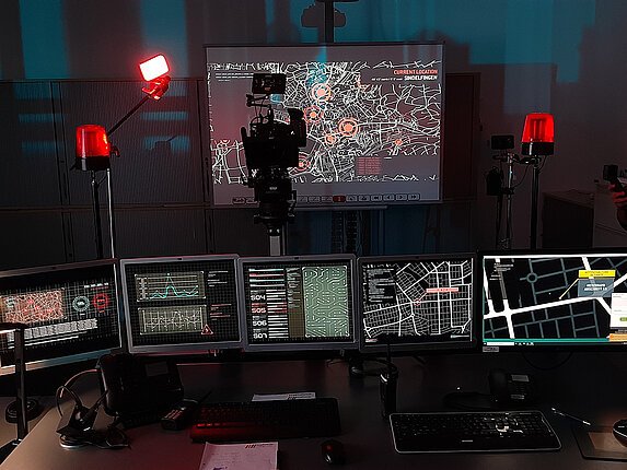 Eine Leinwand mit der Karte von Sindelfingen und einigen roten Markierungen auf dieser Karte. Davor steht eine Filmkamera, die diese Karte aufnimmt. Im Vordergrund ein Arbeitsplatz mit weiteren Bildschirmen und diversen Karten, Analysen und Übersichten.