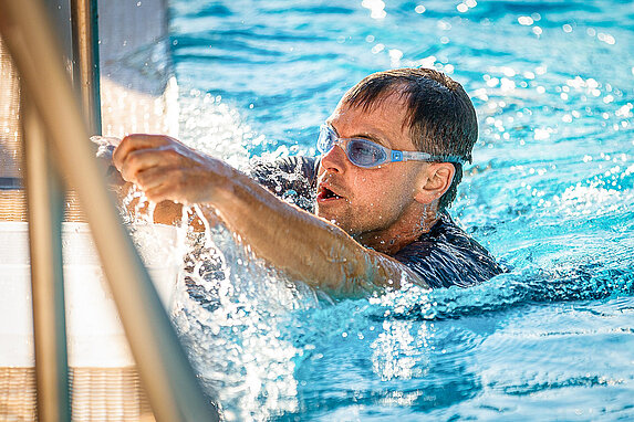 Nahaufnahme eines Schwimmers mit Schwimmbrille im Wasser. Er kommt gerade am Beckenrand an und taucht auf.