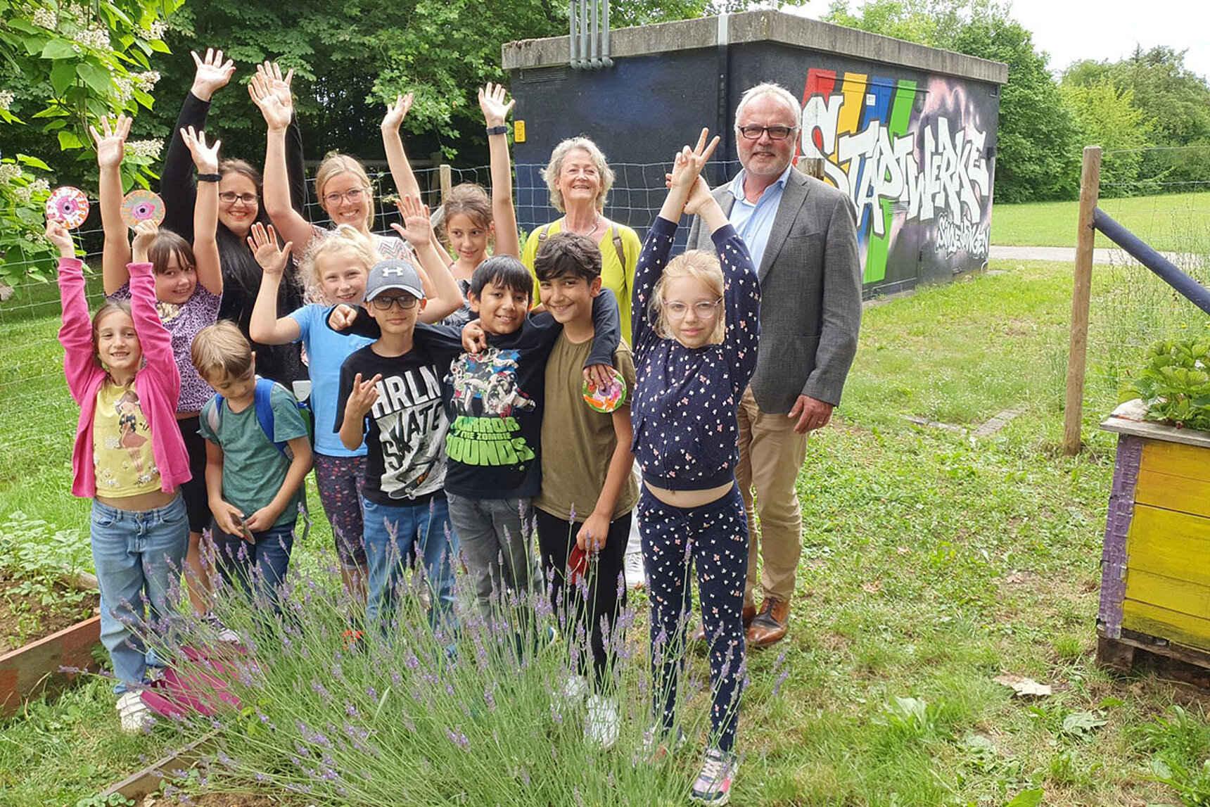 Herr Dr. Hoffmann und einige Kinder stehen zusammen im Projektgarten mit Hochbeeten und Kräutern. Die Kinder freuen sich und strecken die Hände in die Luft.