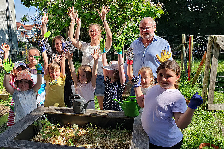 Eine Gruppe Kinder und Herr Hoffmann sind im Schulgarten und stehen um ein Hochbeet herum. Die Kinder heben die Hände in die Luft und freuen sich.