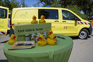 Ein grüner runder Stehtisch auf dem sich einige gelbe Quietscheentchen befinden, dazu ein Schild mit der Aufschrift "Viel Spaß beim Gewinnspiel. Im Hintergrund steht ein Firmenwaagen der Stadtwerke Sindelfingen.