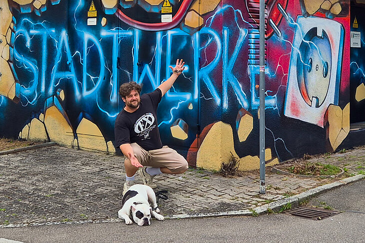 Graffiti-Künstler Max Frank sitzt in der Hocke und mit Hund vor der Station der Stadtwerke. Auf dem Gebäude ist Stadtwerke Sindelfingen als Graffiti gesprüht