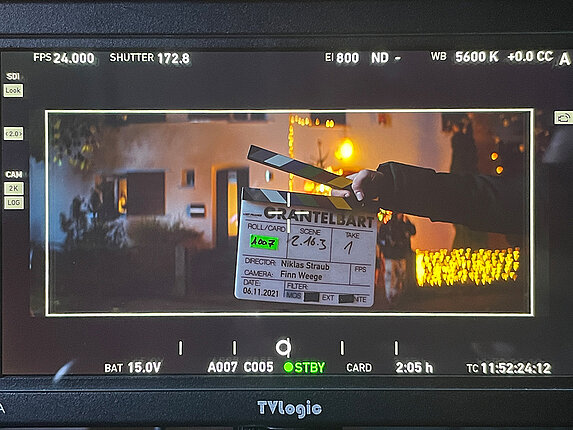 Nahaufnahme eines Displays der Filmkamera. Diverse Anzeigen und Einstellungen rund um die eigentliche Aufnahme. Die Aufnahme selbst zeigt ein Haus, davor eine Hand mit der Filmklappe.