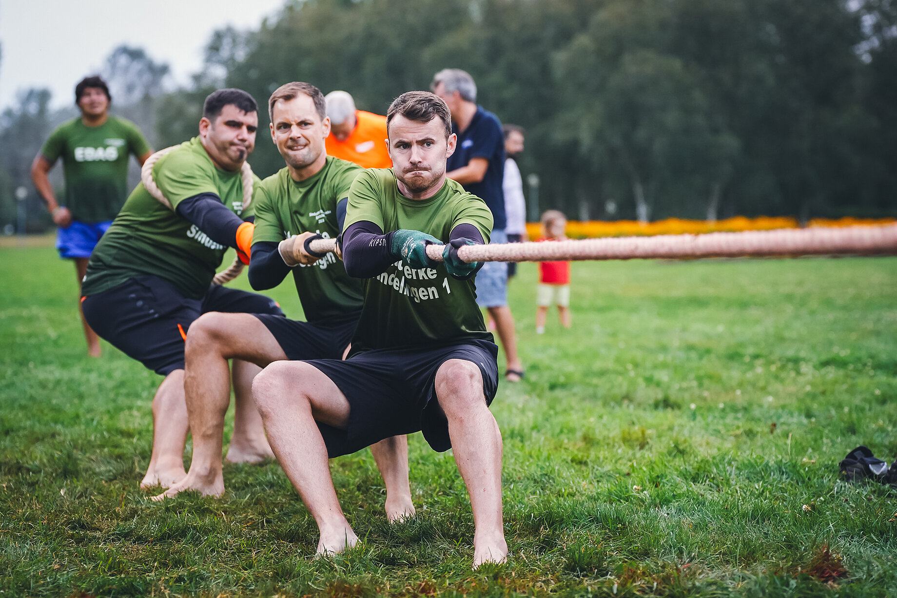 Drei Männer, allesamt barfuß und in Teamkleidung, beim Tauziehen auf einer Wiese. Sie beißen die Zähne zusammen und strengen sich sichtlich an. Im Hintergrund stehen weitere Personen und feuern sie an.