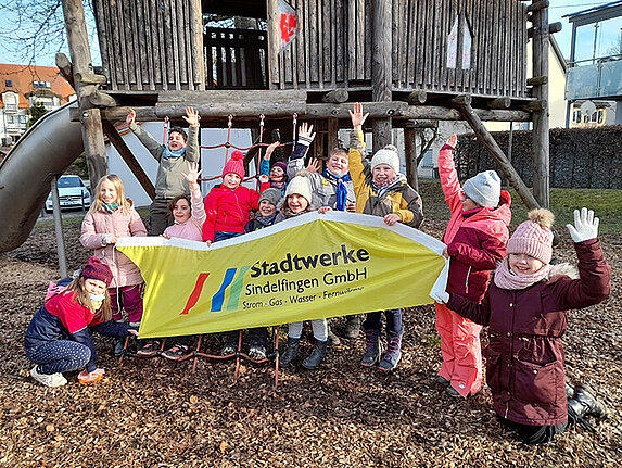 Eine Gruppe Kleinkinder auf einem Spielplatz. Sie halten ein Banner der Stadtwerke Sindelfingen hoch und strecken ihre Hände zur Freude in die Höhe.