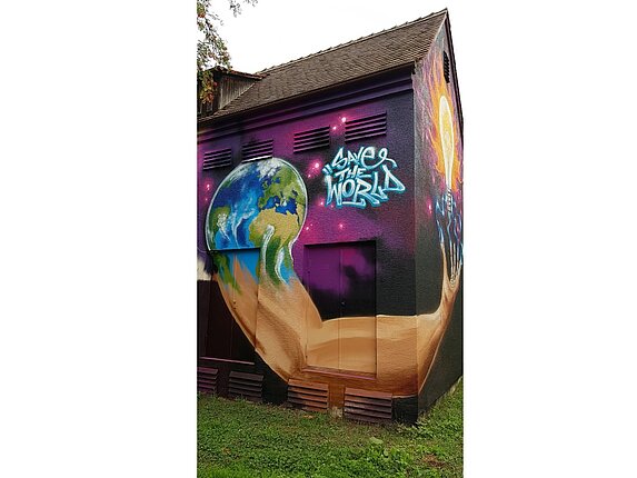 Ein Haus mit einem großflächigen Graffiti. Motiv des Graffitis dieser Seitenwand: Aus der Erdkugel wächst ein Arm heraus. Die Worte "Save the World" und "Save Energy" stehen neben der Erdkugel.