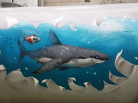 Ein Graffiti auf einem Öltank. Das Motiv: Es scheint als wäre da ein riesiger Riss im Tank, die Ränder sind ausgefranst und mann kann ins Ineere schauen. Dort schwimmt ein grinsender Hai und ein kleiner Fisch in den Farben der Stadtwerke Sindelfingen in hellblauem Wasser. 